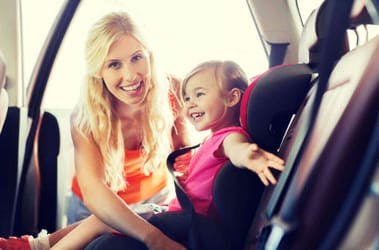 A woman smiling as she straps a small girl into a red child's car seat in the back of a car.  