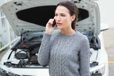 A woman on her phone in front of a car suffering from a breakdown, with the bonnet open. 