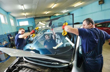 Three mechanics in blue overalls changing the windscreen of a silver car.