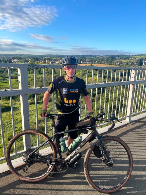 Ollie stood behind his bike on a bridge with the English countryside stretching out behind him