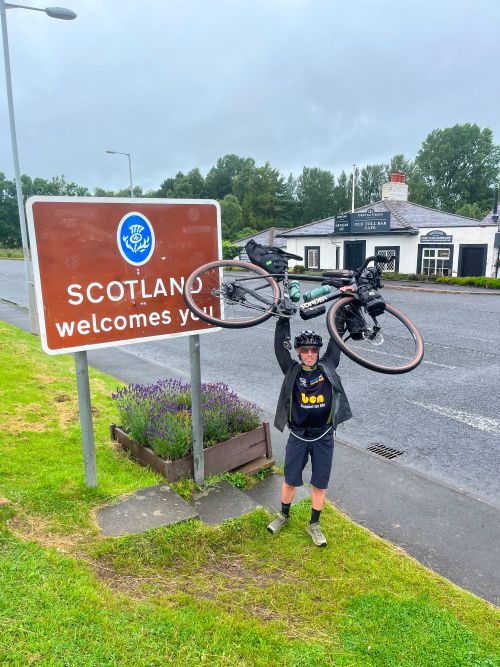 Ollie holding his bike above his head at the Scottish Border during his LEJOG journey